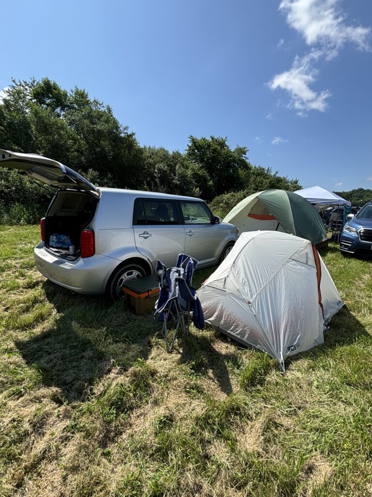 Car with an open trunk. White tent next to the car. Parked on grass at a park. 