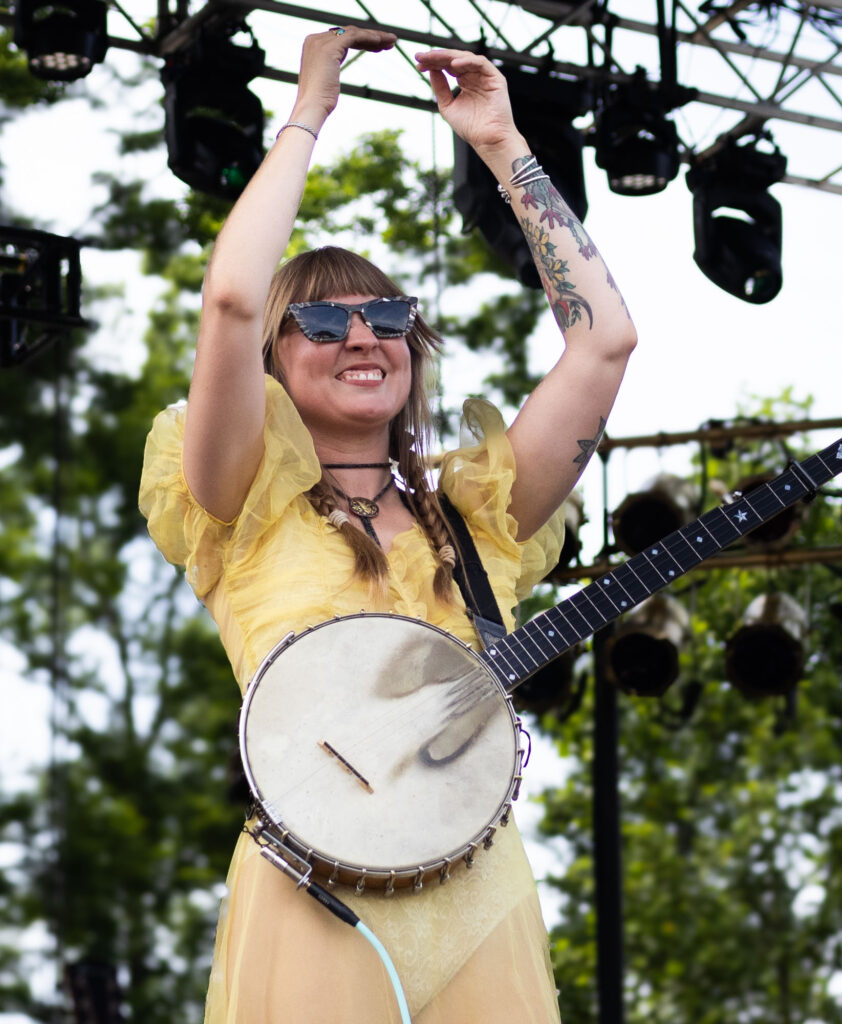 Woman clapping, wearing sunglasses with a banjo slung over her shoulder