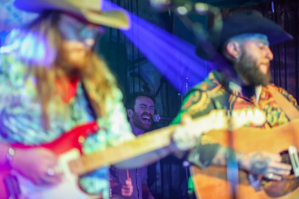 Two people playing guitar with purple lights shining on them, slightly out of focus. Behind them is a drummer singing into a microphone.